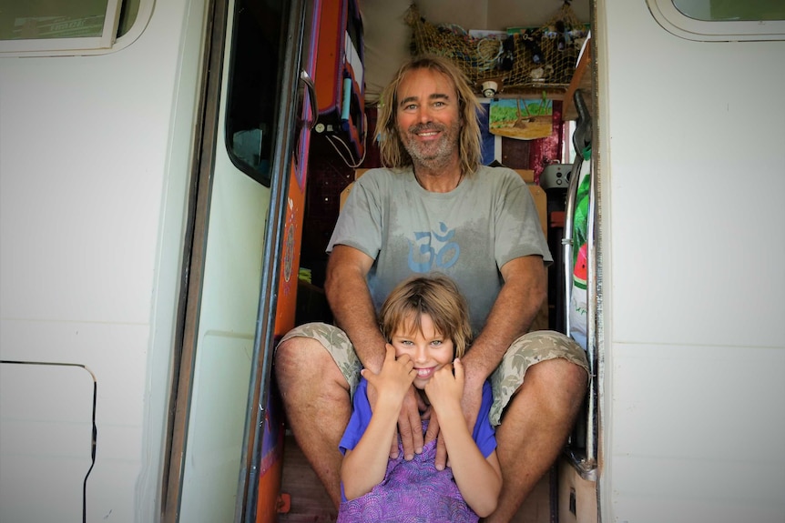 Man hugging daughter sitting on steps of a minibus. Smiling. White bus. Homely interior behind.