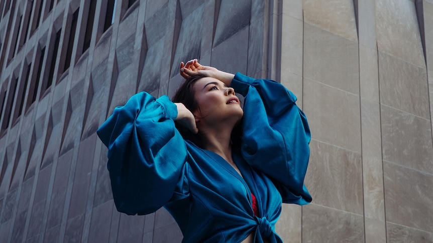 Mitski stands in front of a concrete building, looking up at the sky. She wears a blue dress with big sleeves, tied at the front