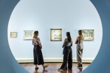 Three young women stand in front of a series of Clarice Beckett paintings in an exhibition