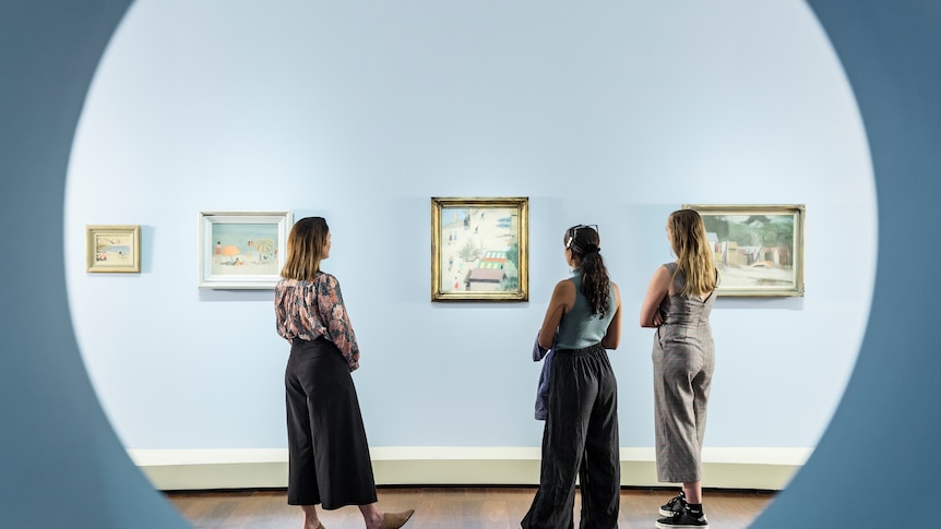 Three young women stand in front of a series of Clarice Beckett paintings in an exhibition