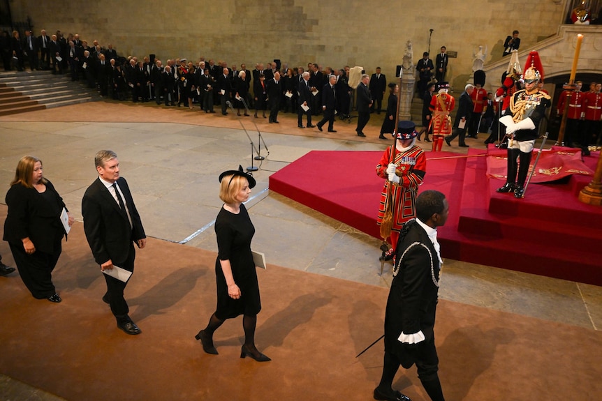 Several men and women dressed in black walk in a procession in a church hall 