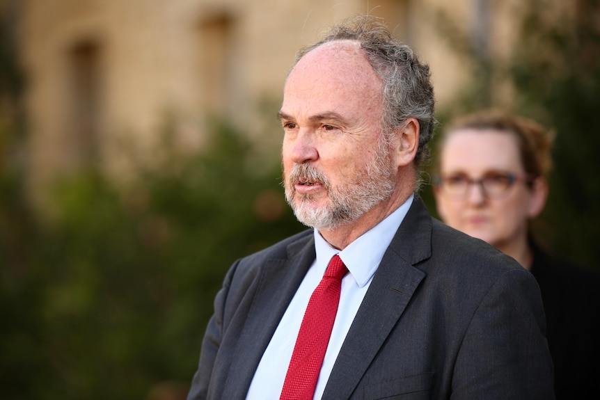 A mid-shot of Bill Johnston speaking at a media conference outside wearing a suit and tie.