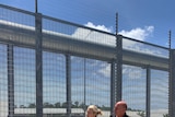 Natasha Griggs points into the distance as she stands alongside a towering fence with barbed wire on top