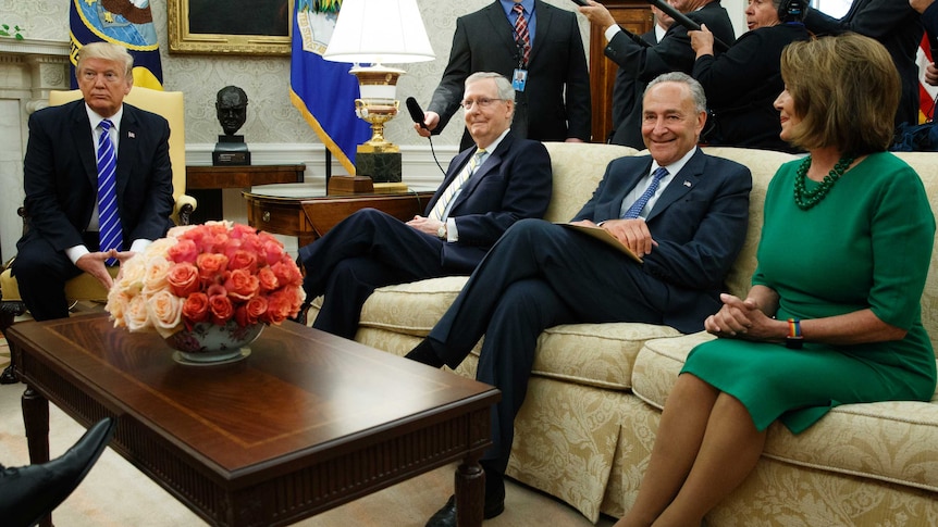 Donald Trump sits at a chair adjacent to a couch, on which Nancy Pelosi, among others, is sitting. Press are in the background.