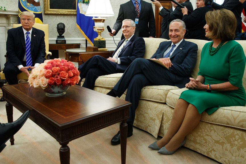 Donald Trump sits at a chair adjacent to a couch, on which Nancy Pelosi, among others, is sitting. Press are in the background.