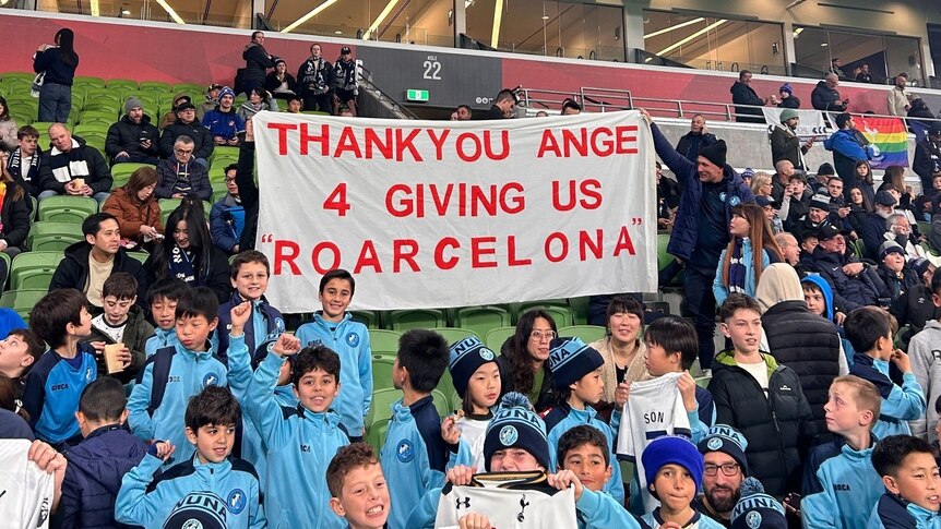 Young soccer fans seated around a sign made for Ange Postecoglou.
