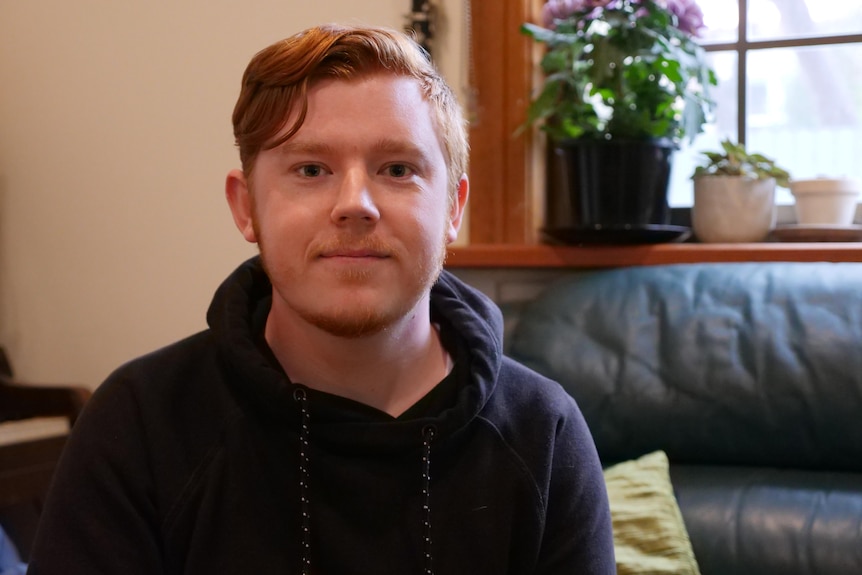 A red haired man wearing a black hoodie sits on a leather couch looking at the camera.