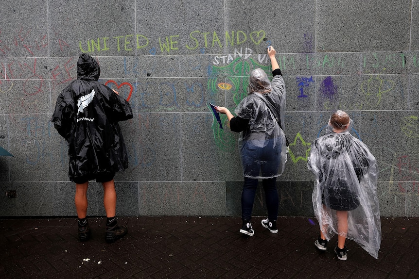 Three men in dark raincoats write messages on soggy wall with chalk including United We Stand