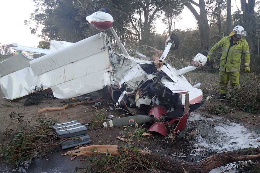 The wreckage of a light plane lies on the ground in bush with a firefighter standing alongside the aircraft.