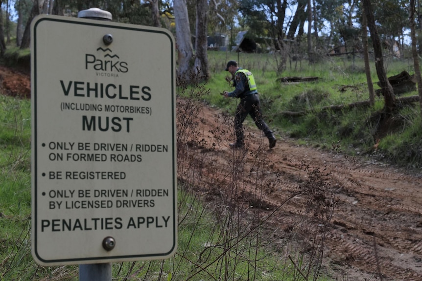 A sign tells drivers to stay off the track, muddy track, a man in uniform with a high viz walking on track, trees, grass.