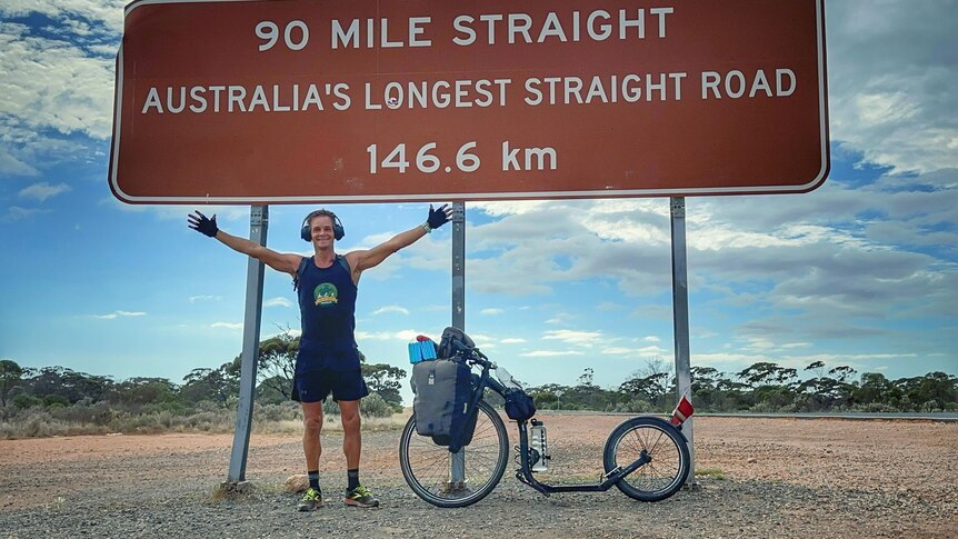 Smiling man, arms spread out, stands underneath road sign 90 Mile Straight Australia's Longest Straight Road 146.6km, kick scoot
