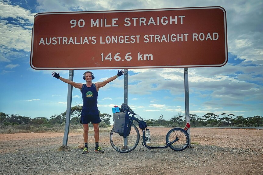 Man with cycle stands beneath sign saying it's Australia's longest straight road