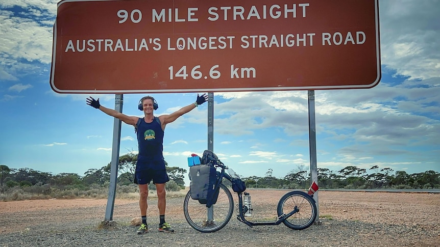 Smiling man, arms spread out, stands underneath road sign 90 Mile Straight Australia's Longest Straight Road 146.6km, kick scoot