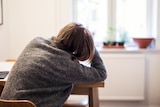 A person in a grey sweater slumps at a kitchen table.