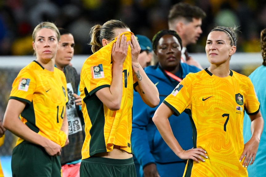 Three Australian female football players look upset, one covers her face, after defeat in a World Cup match.