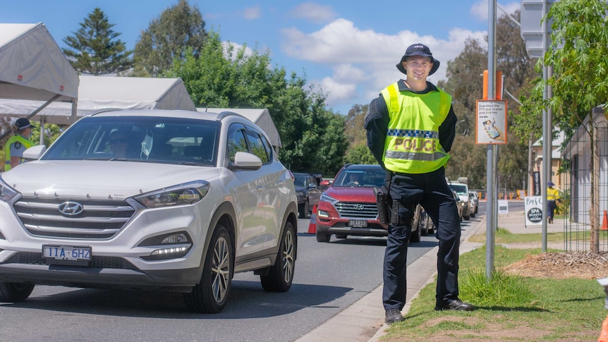 Man in police uniform stands on side of road next to cars passing tents at border checkpoint