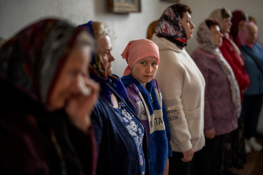 A young girl stands among a group of older women all wearing headcoverings inside a church. 