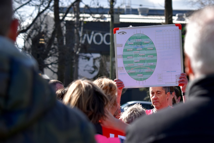 A man holds a whiteboard with a picture of an Aussie Rules Oval and positional markers