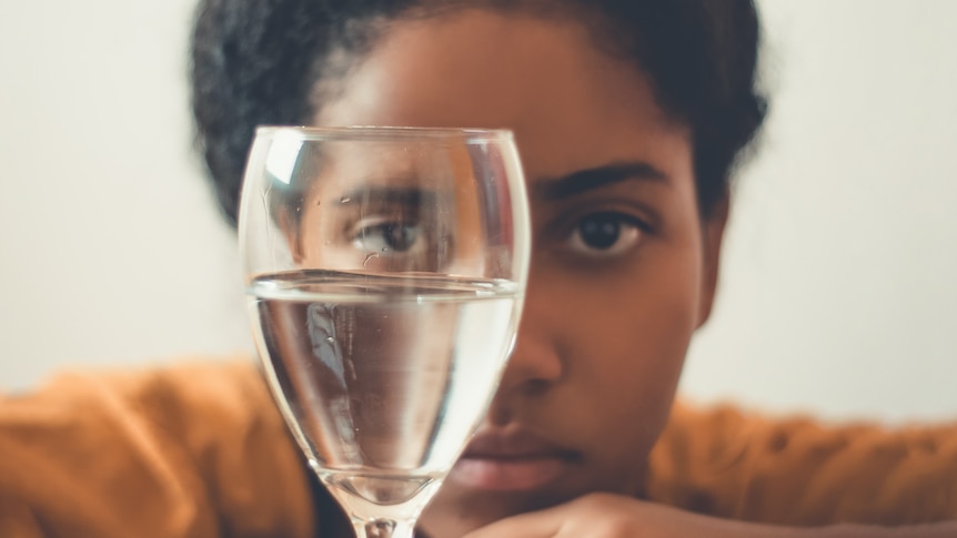 A woman rests her chin on a table and looks at a wine glass half full of water