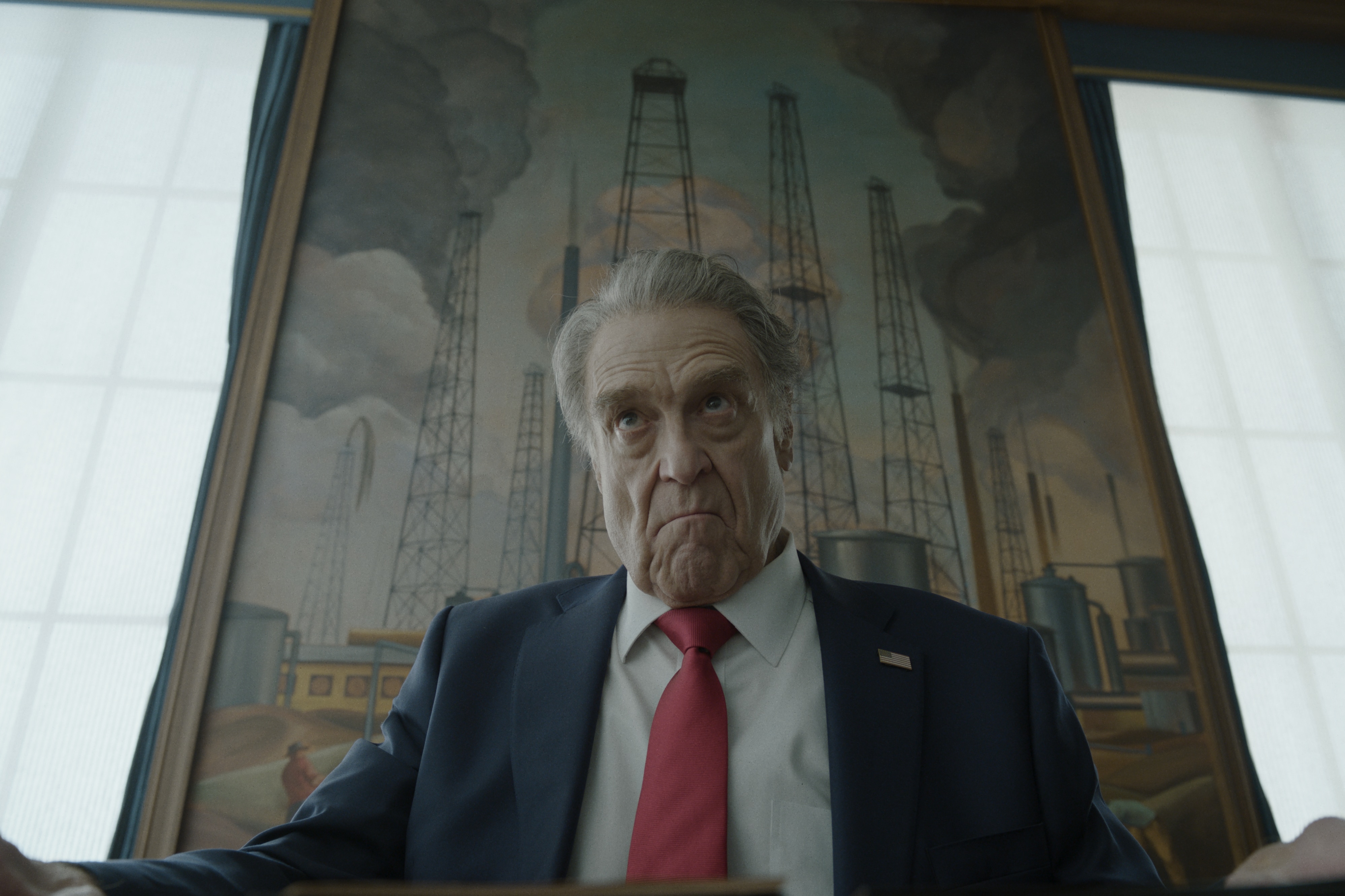 A suited man (John Goodman) sits at a table with an angry face.