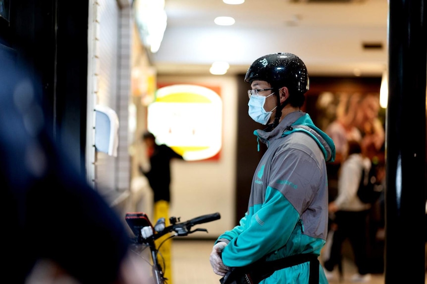 A food delivery man wearing a black helmet and a teal jacket stands in front of a fast food restaurant window.