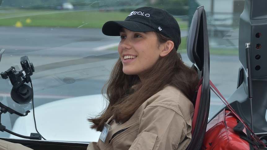 A portrait of a young woman with long brown hair sitting in a small microlight aircraft