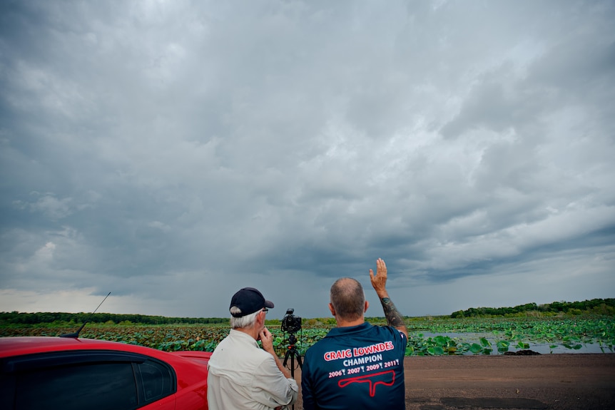 The backs of two men, with a camera inbetween, looking at a stormy sky. 