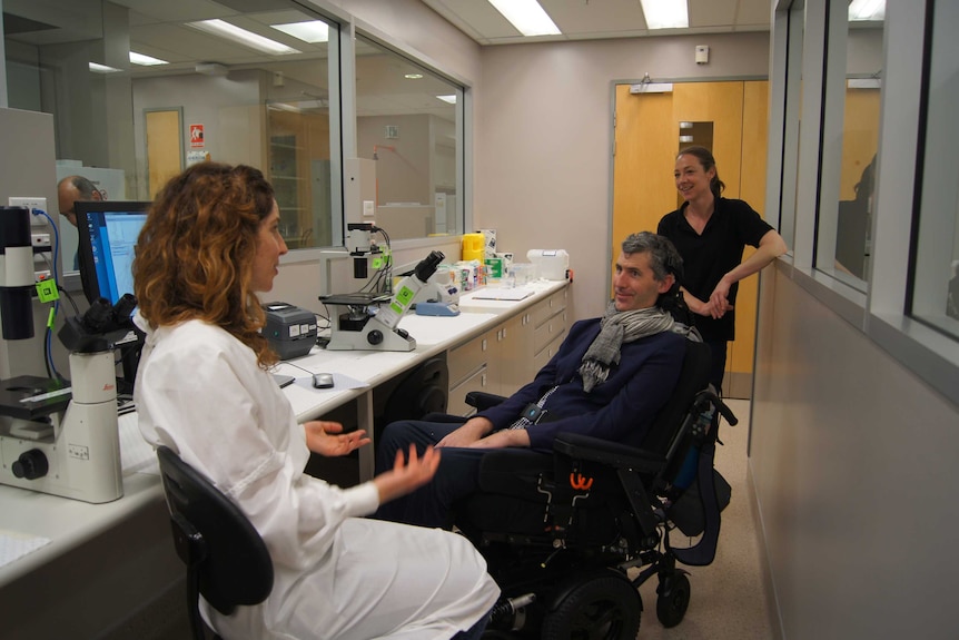 A man in a wheelchair speaks with a woman in a lab coat while another woman watches on.