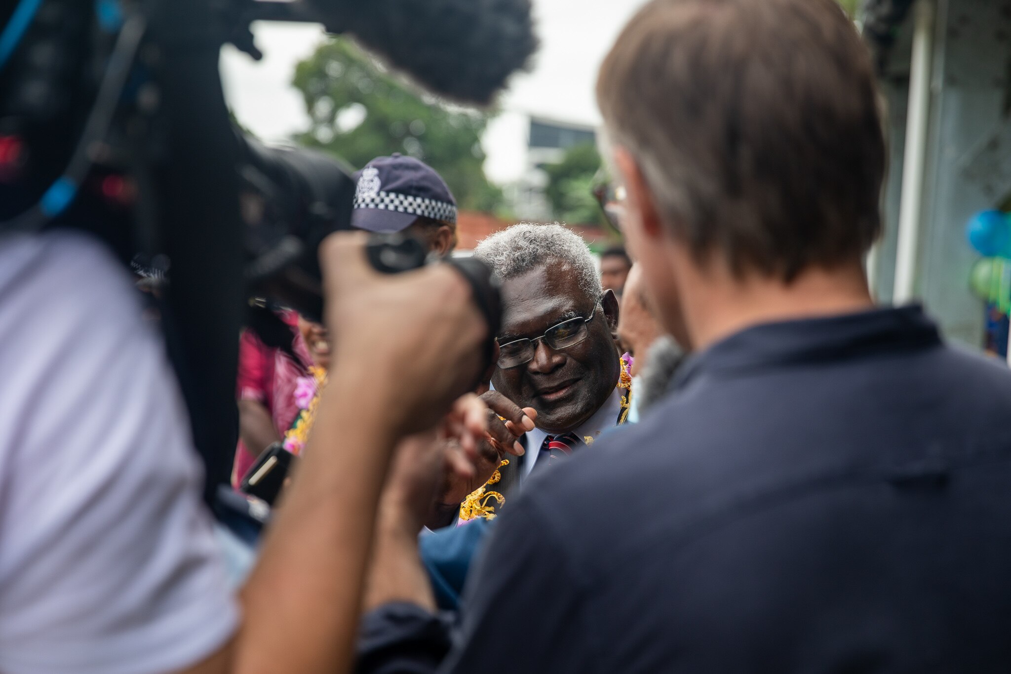 The Prime Minister holds up a finger. He's surrounded by people, including in the foreground and journalist and camera operator.