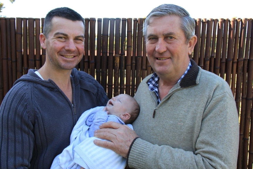A family photo of an older man with grey hair, his middle aged son who holds his newborn in backyard