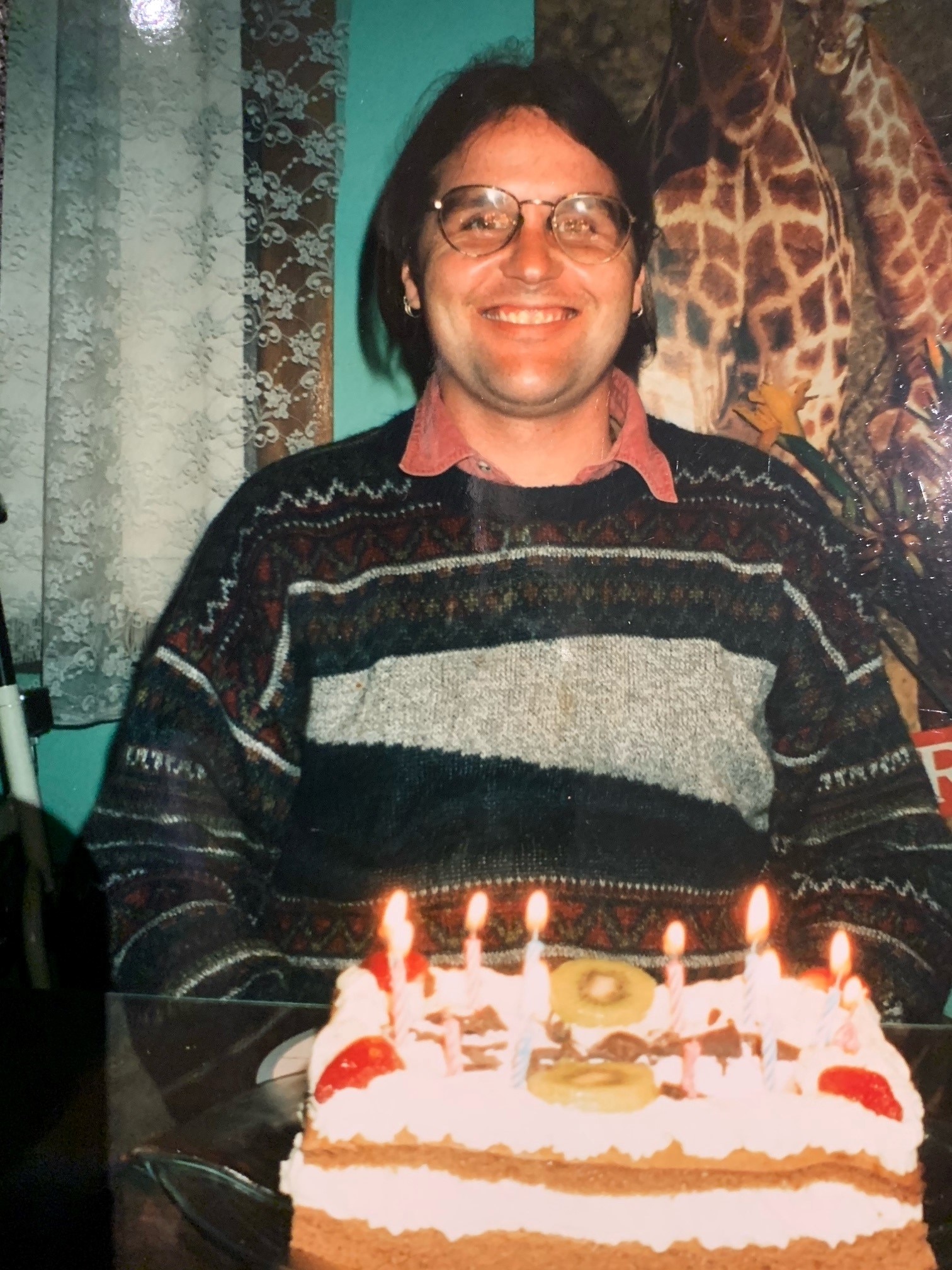 A man wearing a jumper and glasses smiles and sits behind a large birthday cake.