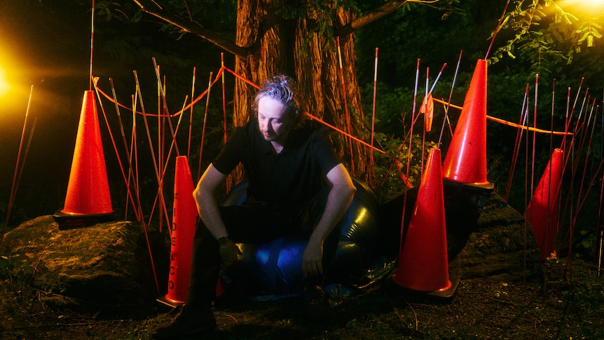 Daniel Lopatin of OPN is photographed sitting under a tree in the dark surrounded by red witches hats