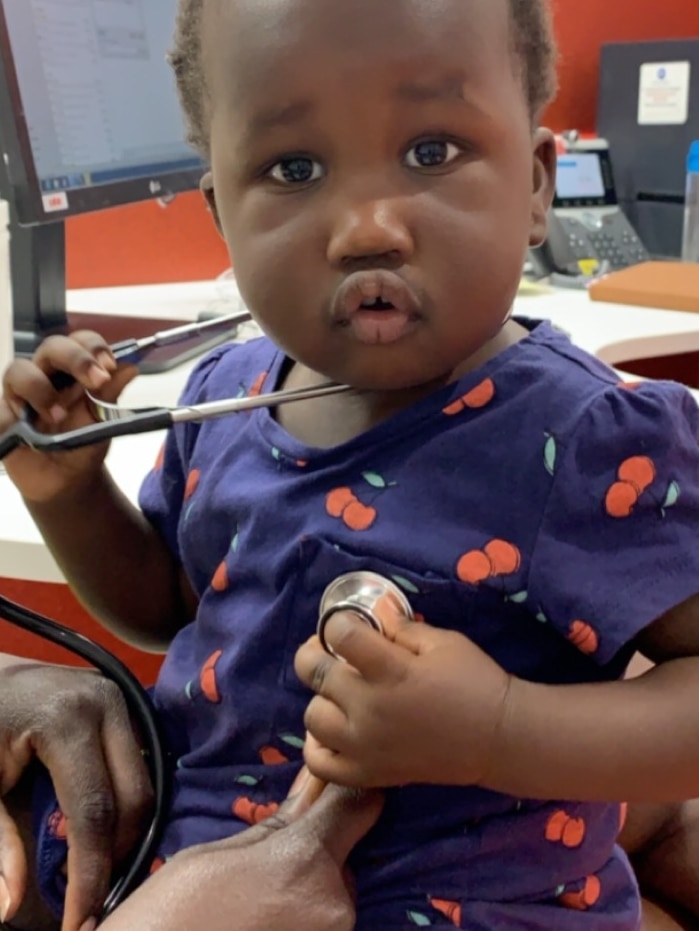 Nyankur Bol sitting on someone's lap with a stethoscope around her neck and on her chest.