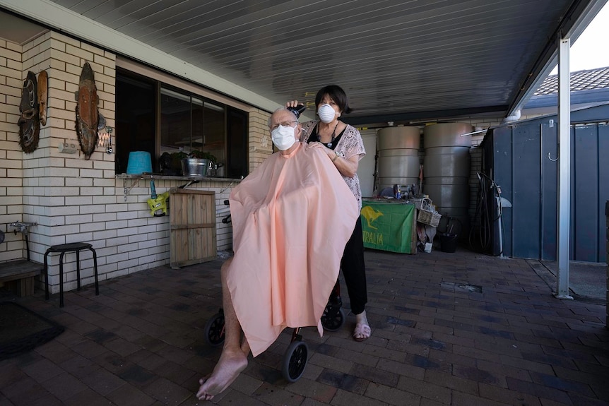 A woman wearing a facemask cuts a man's hair in the garage of their home. He wears a facemask too.