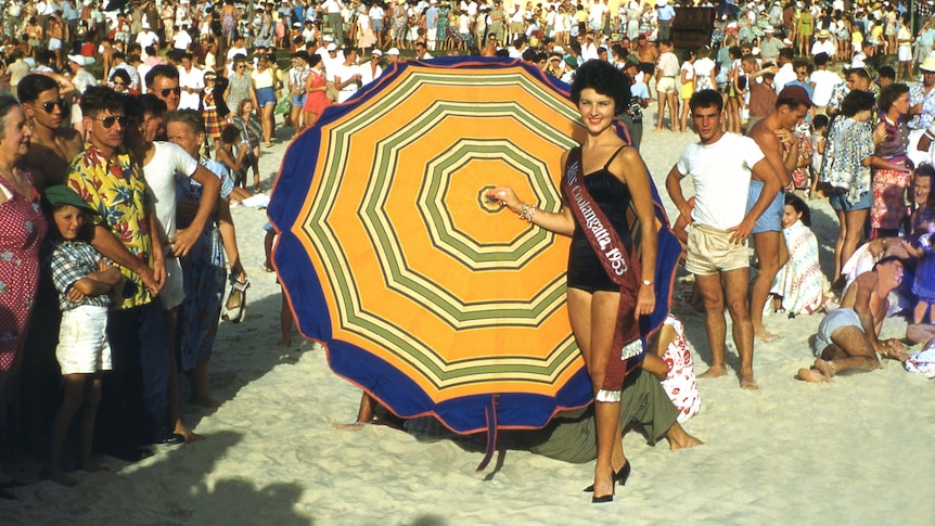 A woman wearing a black one-piece swim suit stands in front of a large striped umbrella on a crowded sandy beach.