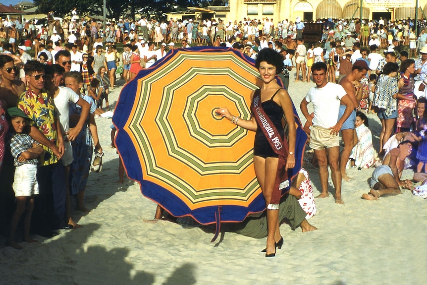 A woman wearing a black one piece swim suit stands in front of a large stripped umbrella on a crowded sandy beach 