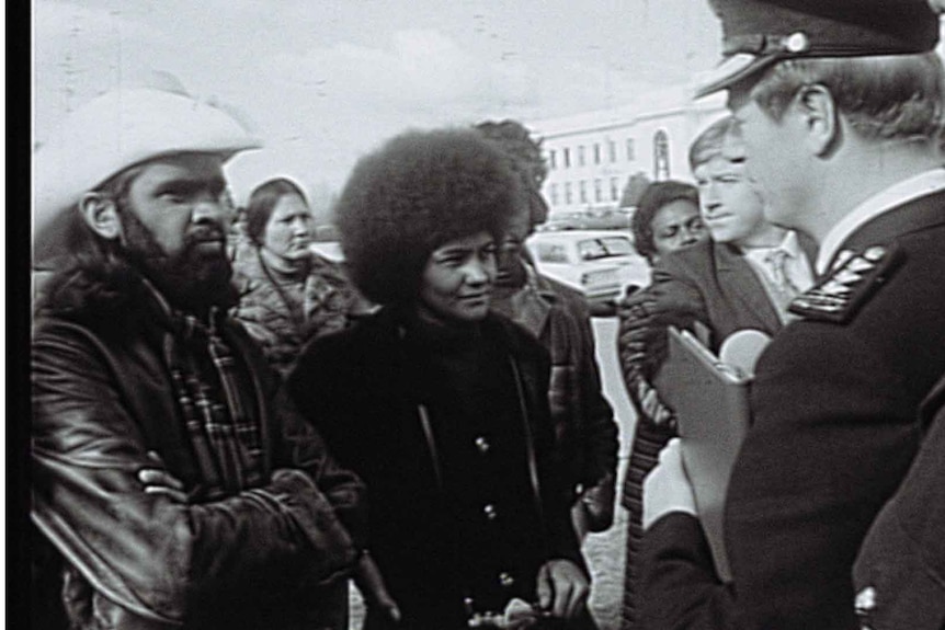 A woman with an Afro surrounded by a small group of people facing uniformed law enforcement