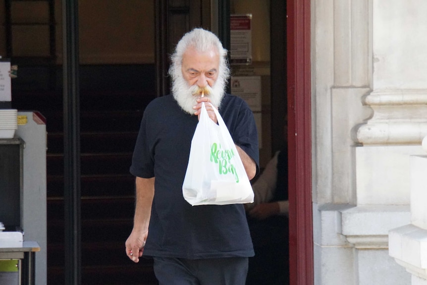 A white-bearded man leaving a building smoking and holding a plastic bag.