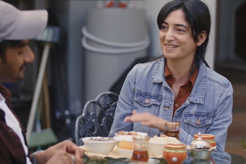 A woman in denim jacket speaking with tea and food on table.