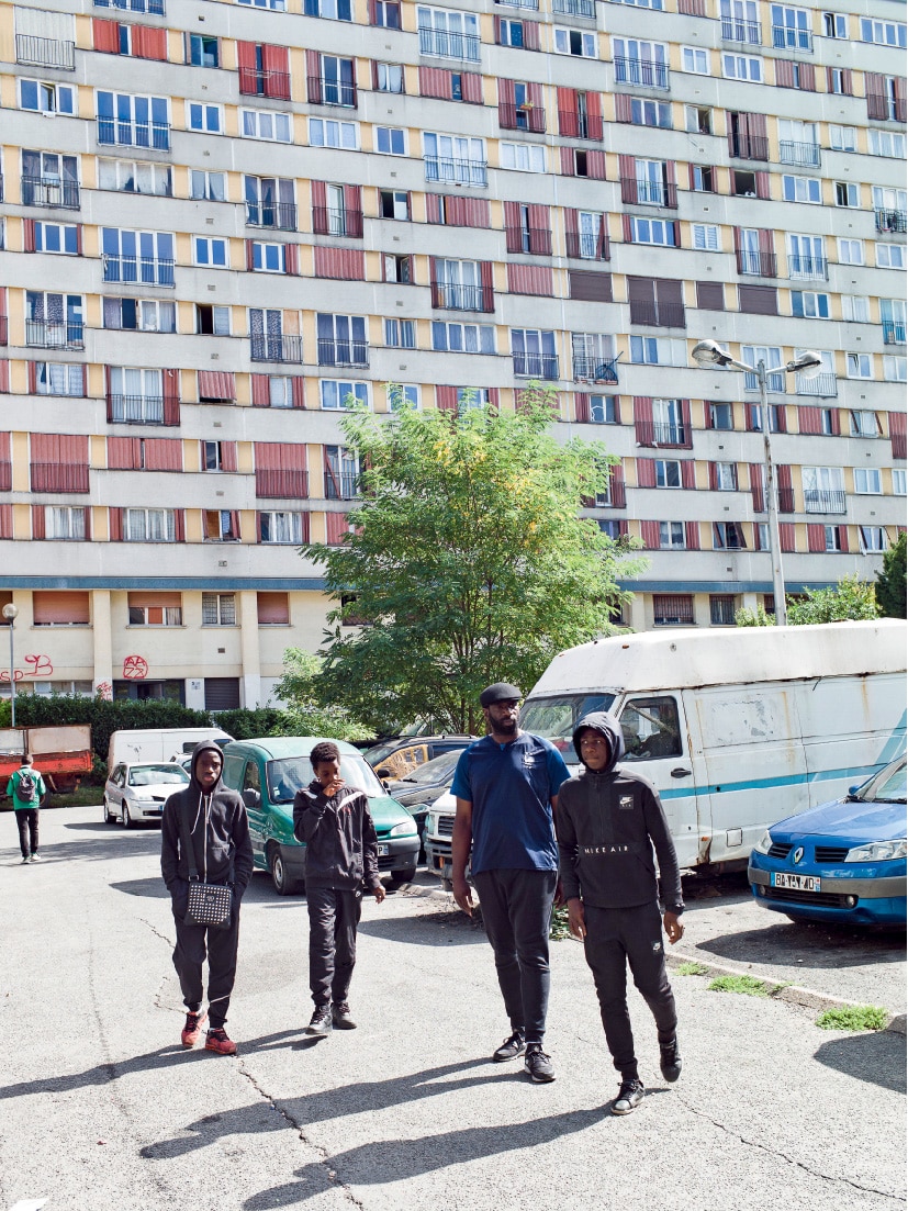 Figures walking in car park, with the gridded facade of a housing project behind them.