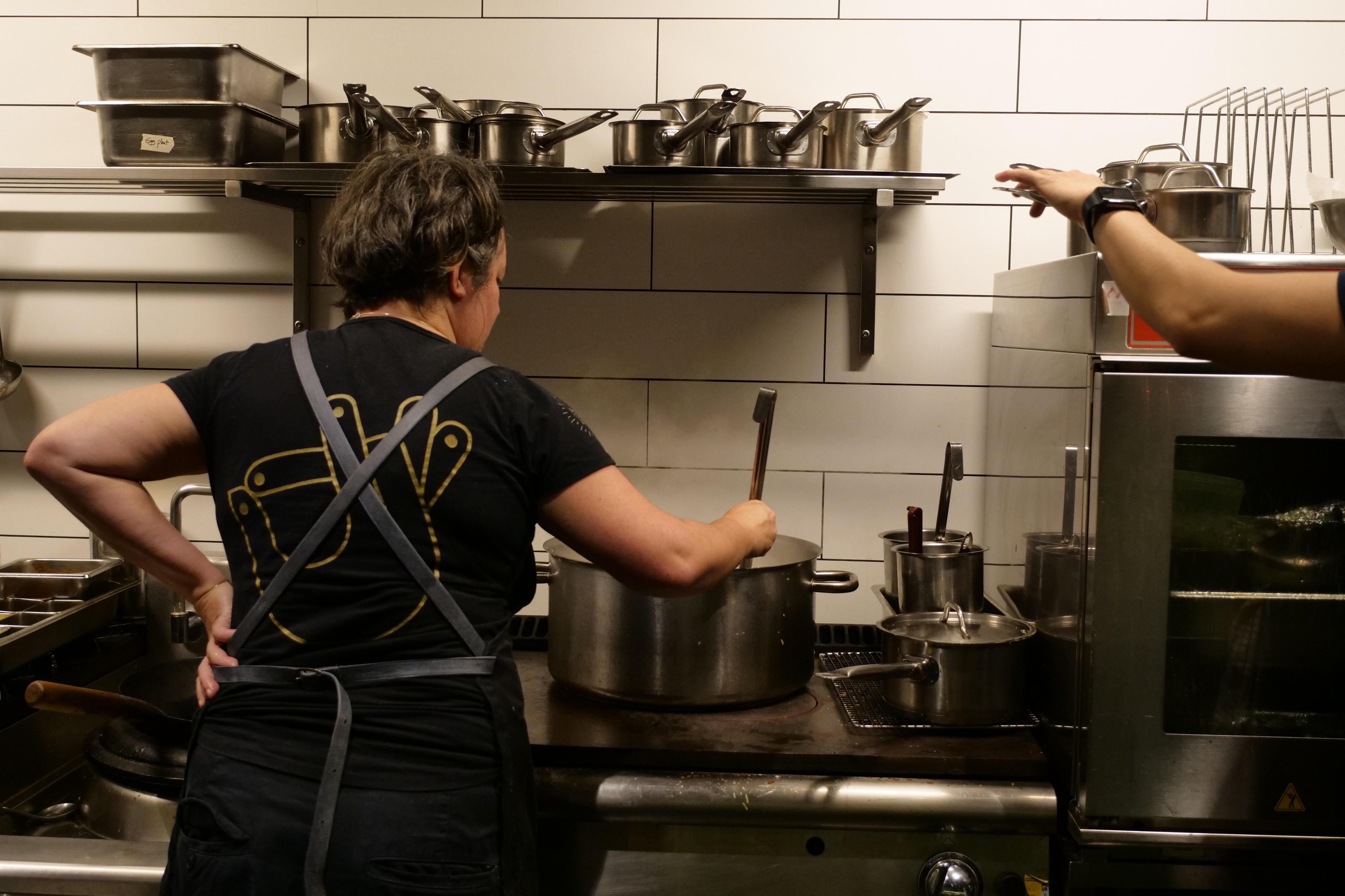A woman with black hair in a black t-shirt is stirring a large pot in the kitchen