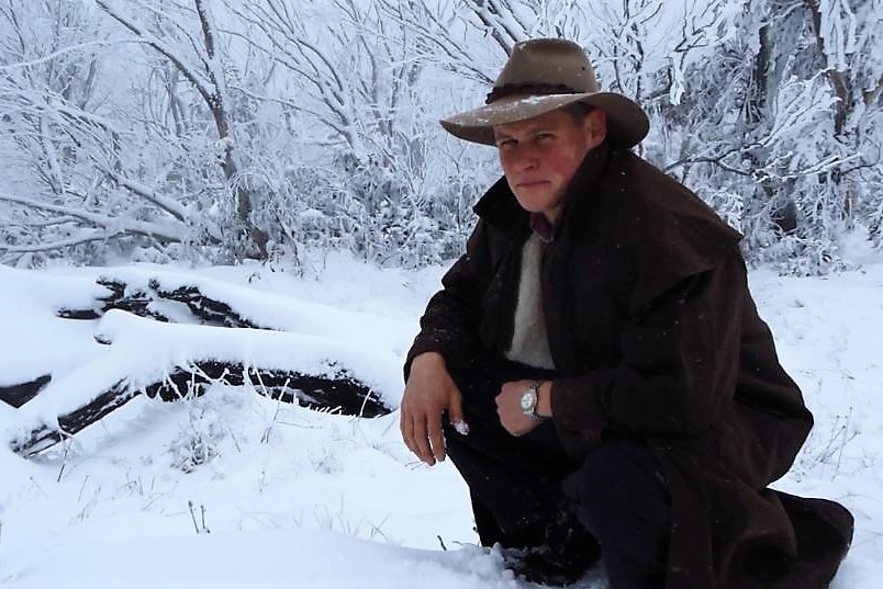Bill Spencer crouches in the snow, wearing a long coat.