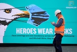 A construction worker wearing a mask passes a billboard that reads 'heroes wear masks'