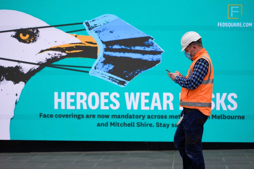 A construction worker wearing a mask passes a billboard that reads 'heroes wear masks'