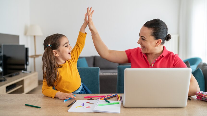 Mother and young daughter sit at a kitchen table working on homework and then high five. 