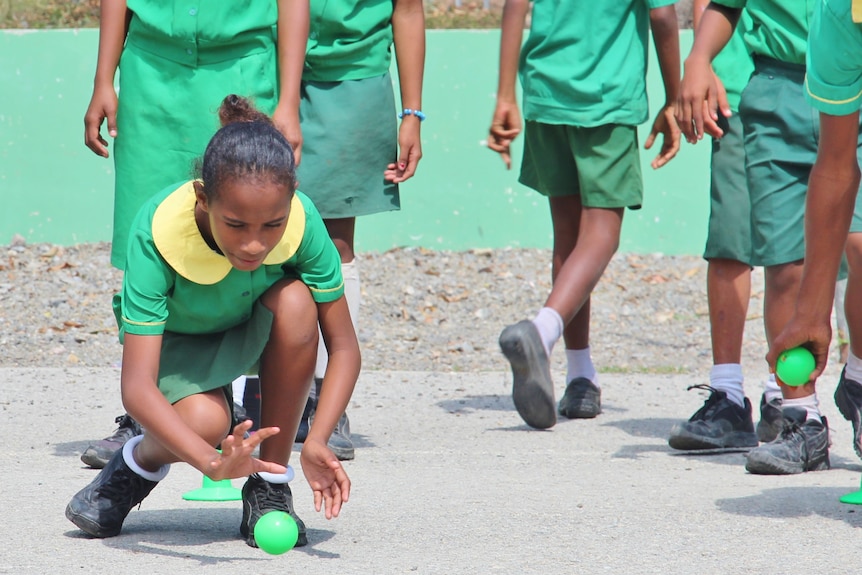 A girl in a green school uniform crouches to pick up a green ball travelling towards her.