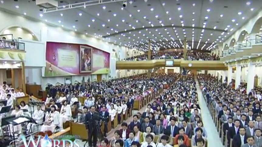 A large auditorium hosts a crowd of Korean followers of the Manmim Central Church with camera operators in foreground