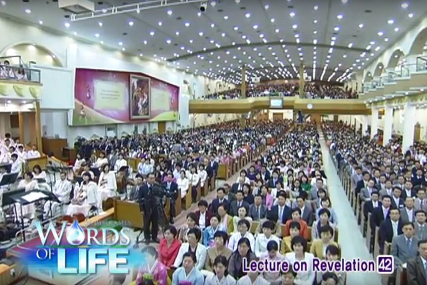 A large auditorium hosts a crowd of Korean followers of the Manmim Central Church with camera operators in foreground