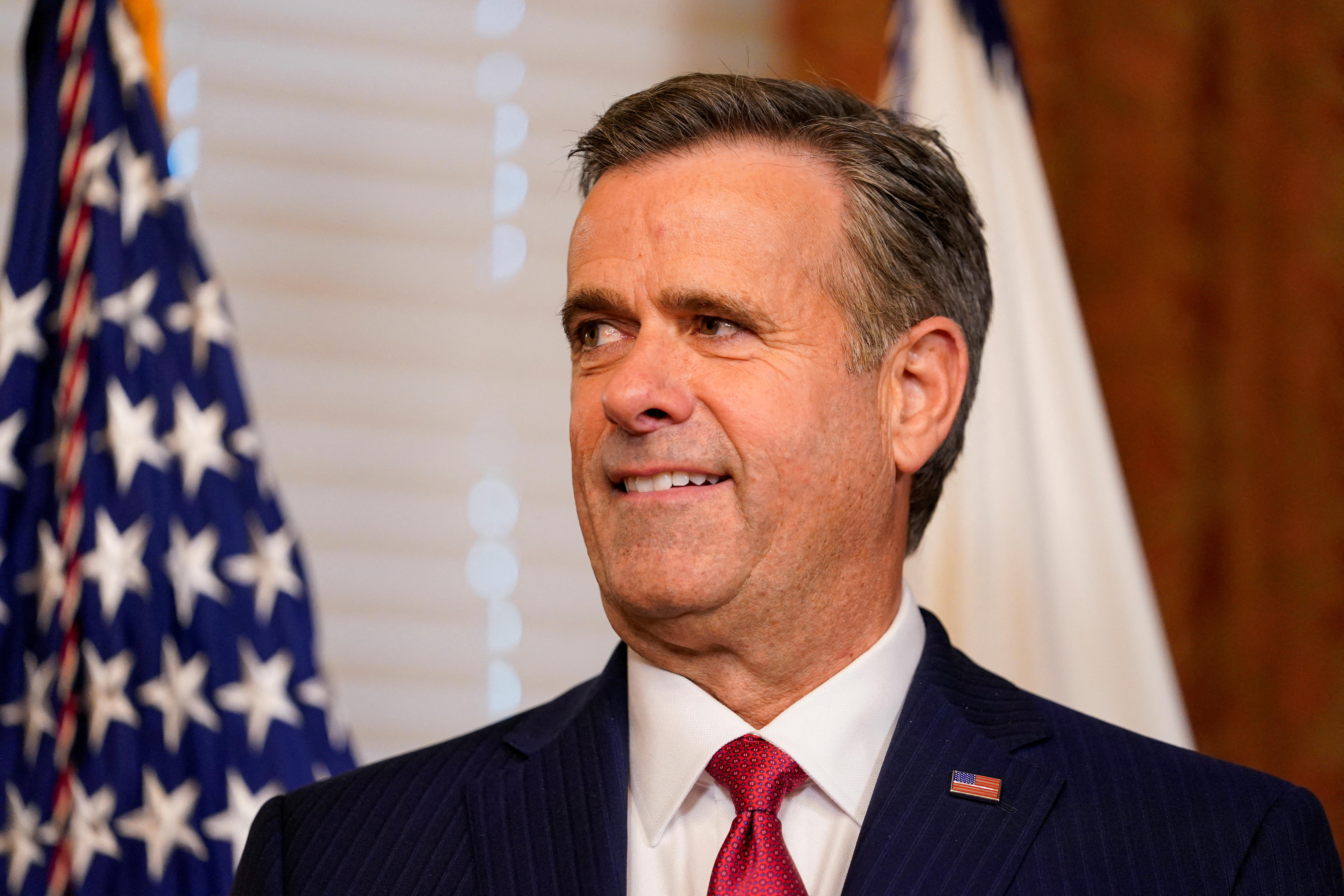 Ratcliffe stands in an office with venetian blinds and a US flag behind him 