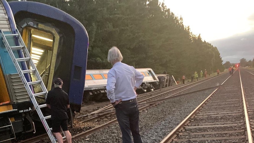A train lies on its side, a man with his hands on his hips looks at the carriage on its side.